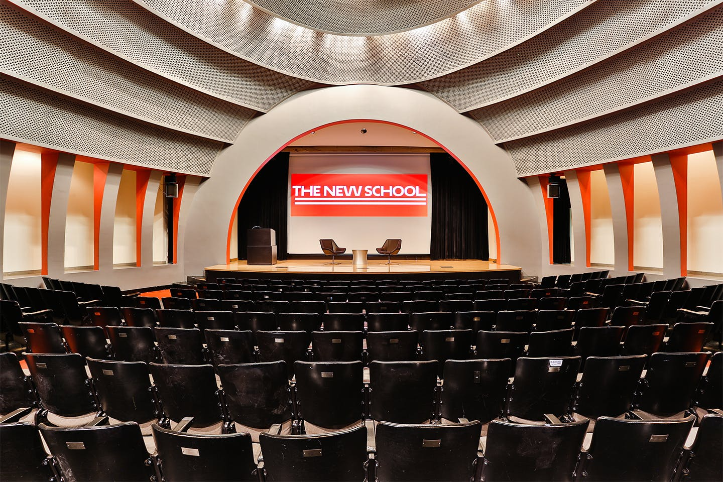 Large auditorium with fixed audience seating and dramatic dome made up of concentric circles. The small elevated stage shows a podium, a projection screen behind a curtain, and two chairs for a seated conversation.